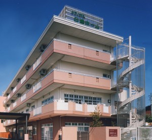 ピンク色の外壁が特徴の介護施設の建物外観。複数階建てで階段室が見える。